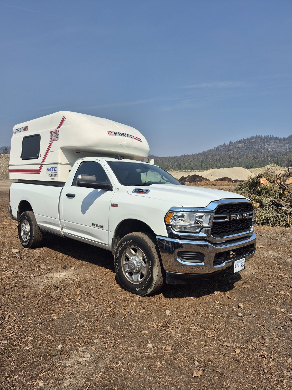 White Ram 3500 truck carrying a First Aid Mobile Treatment Centre unit, parked at the wildfire cleanup site in Summerland, BC, with piles of cleared brush and fire-scorched hillsides behind under a smoke-hazy sky